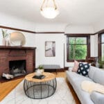 A cozy living room with a gray sofa, round coffee table, and a brick fireplace. A circular mirror hangs above the mantel, and a large window lets in natural light. Decorative pillows and a rug add comfort and style. 