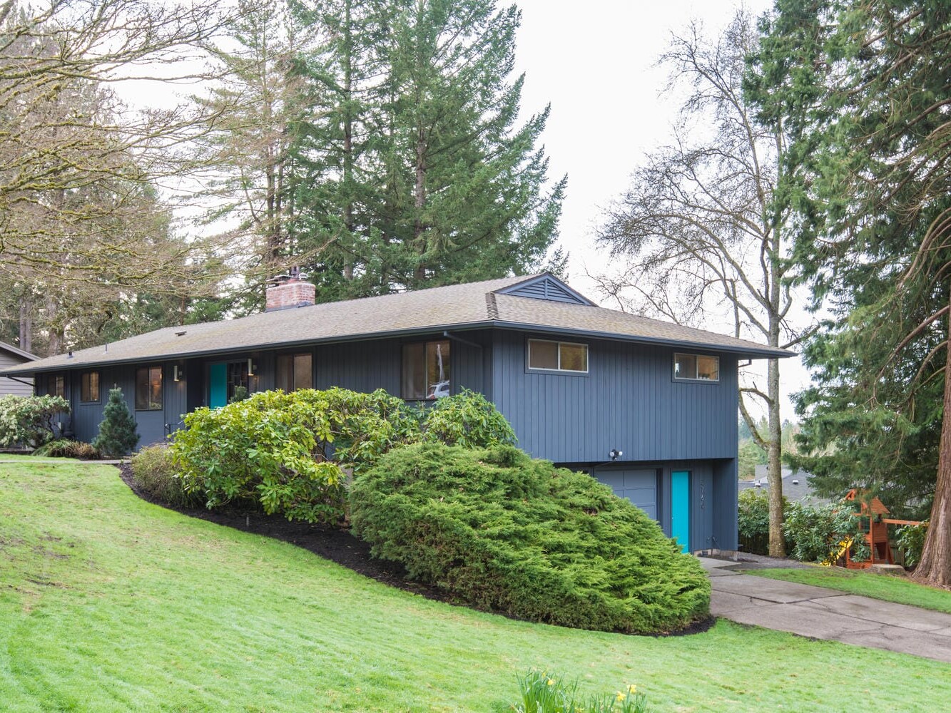 A blue, single-story house with a sloped roof nestles among tall trees in Portland, Oregon. The front yard features a manicured lawn, shrubs, and a driveway leading to a two-car garage. The overcast sky enhances the tranquil atmosphere.