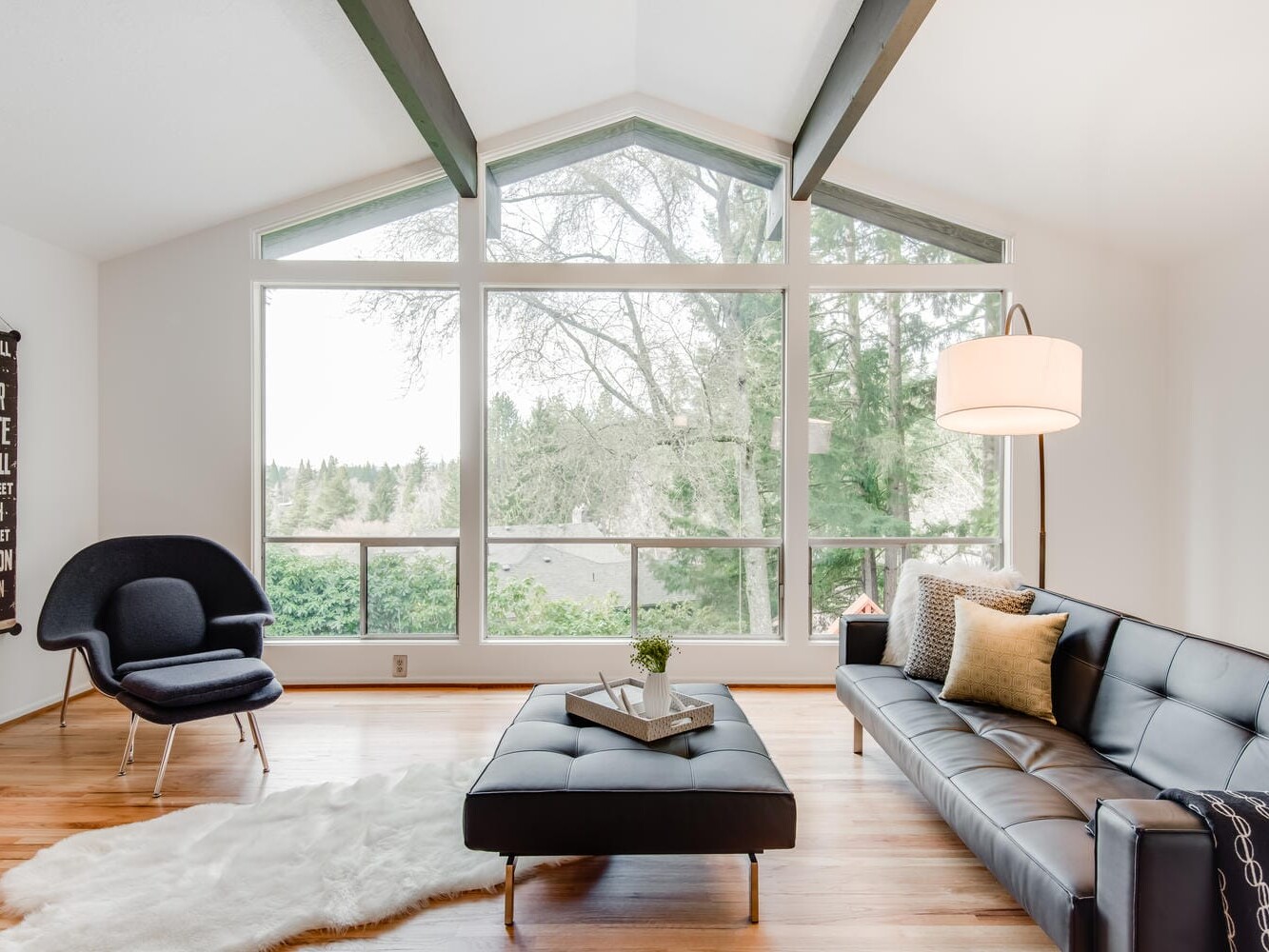A bright, modern living room in Portland, Oregon, boasts a large window showcasing trees outside. It features a black leather sofa, a black armchair, a central ottoman with tray, a white fur rug, and a floor lamp. A decorative sign graces one wall.