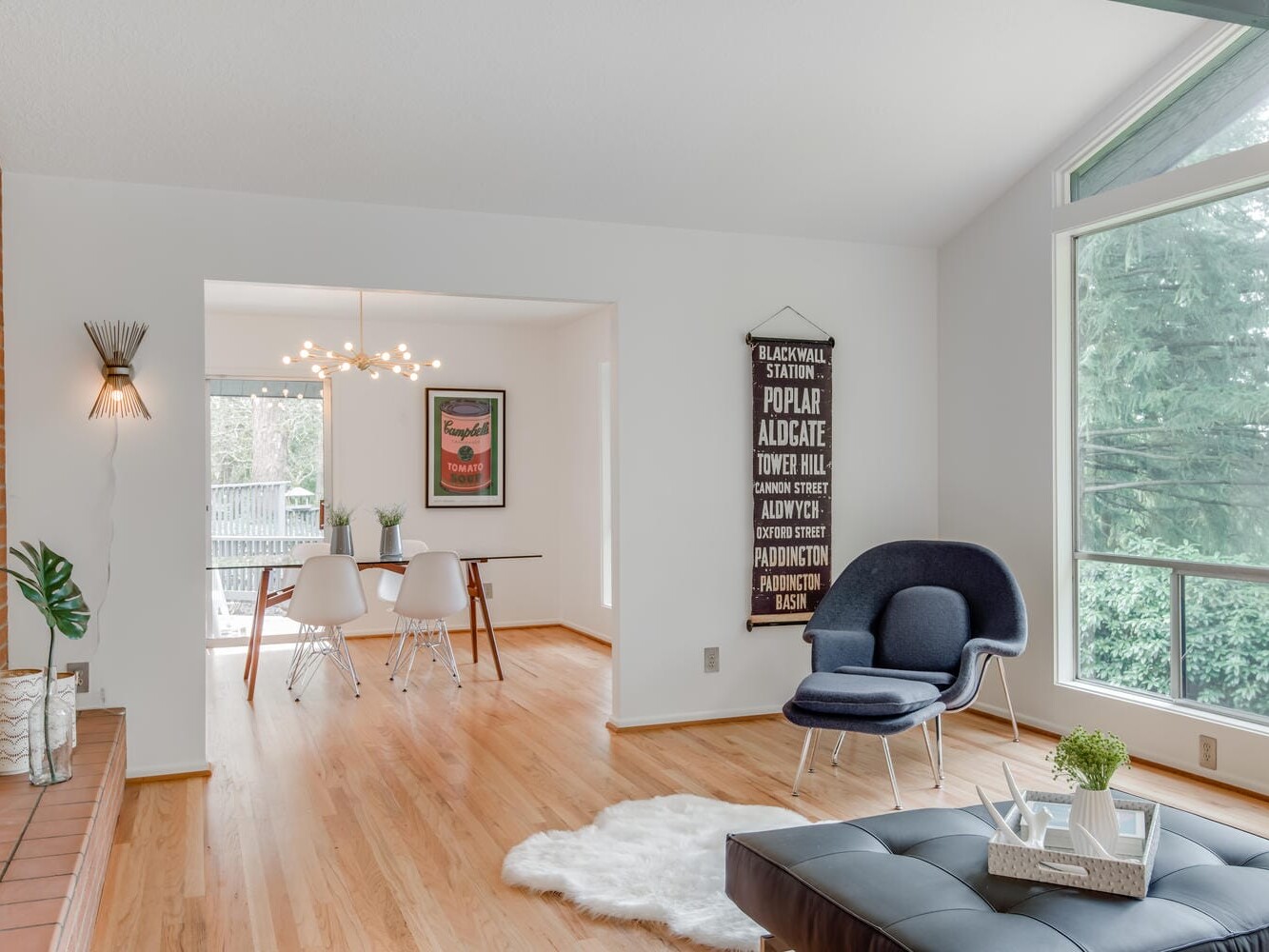 A modern living room in Portland, Oregon showcases light wood flooring, a blue armchair by a large window, and a white furry rug. The brick accent wall features two plant vases, while the dining area presents a round table with white chairs and a minimalist chandelier.