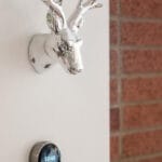 A chrome deer head wall decoration, reminiscent of trendy Portland, Oregon aesthetics, is mounted above a digital thermostat displaying 10:49 on a pristine white wall. A brick wall is partially visible to the right. 