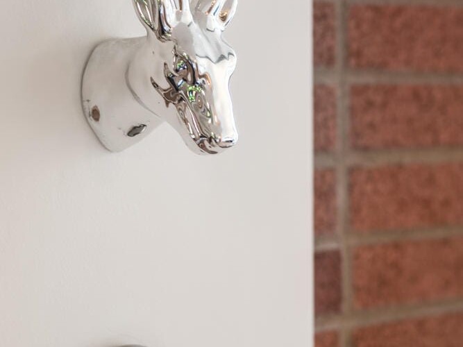 A chrome deer head wall decoration, reminiscent of trendy Portland, Oregon aesthetics, is mounted above a digital thermostat displaying 10:49 on a pristine white wall. A brick wall is partially visible to the right.