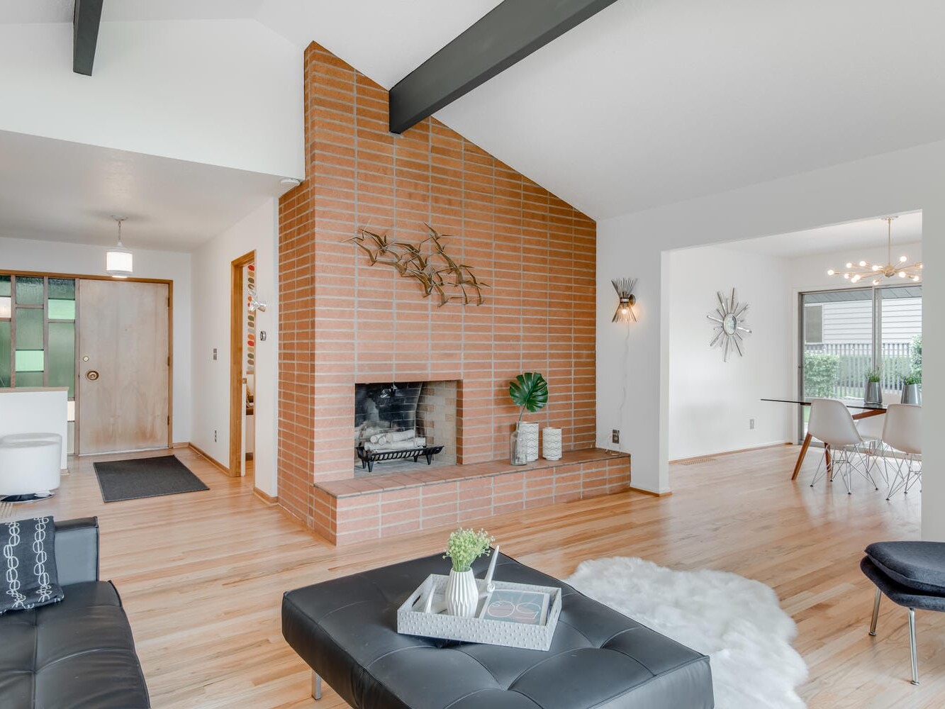A modern living room in Portland, Oregon features a brick fireplace and angled metal decor. The space boasts a black leather ottoman, light wooden floors, white walls, and a high beamed ceiling. In the background, a dining area with a round table and chairs is visible.