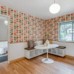 A retro-style dining room reminiscent of Portland, Oregon, with orange and brown leaf-patterned wallpaper. It features a round white table, built-in L-shaped bench seating with cushions, and a window showing an outside view. A round ceiling light softly illuminates the cozy space. 