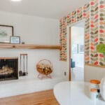 A cozy living room in Portland, Oregon, features a white brick fireplace with a wooden mantel adorned with decorative items. The wall boasts patterned wallpaper with colorful leaf designs. A white table holding a vase and green decorative spheres sits invitingly in the foreground. 