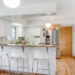 Bright, modern kitchen in Portland, Oregon with a wooden floor, white cabinets, and a stainless steel refrigerator. A patterned backsplash and colorful bowls add charm. Three white bar stools line the kitchen island while a large window fills the space with natural light. 