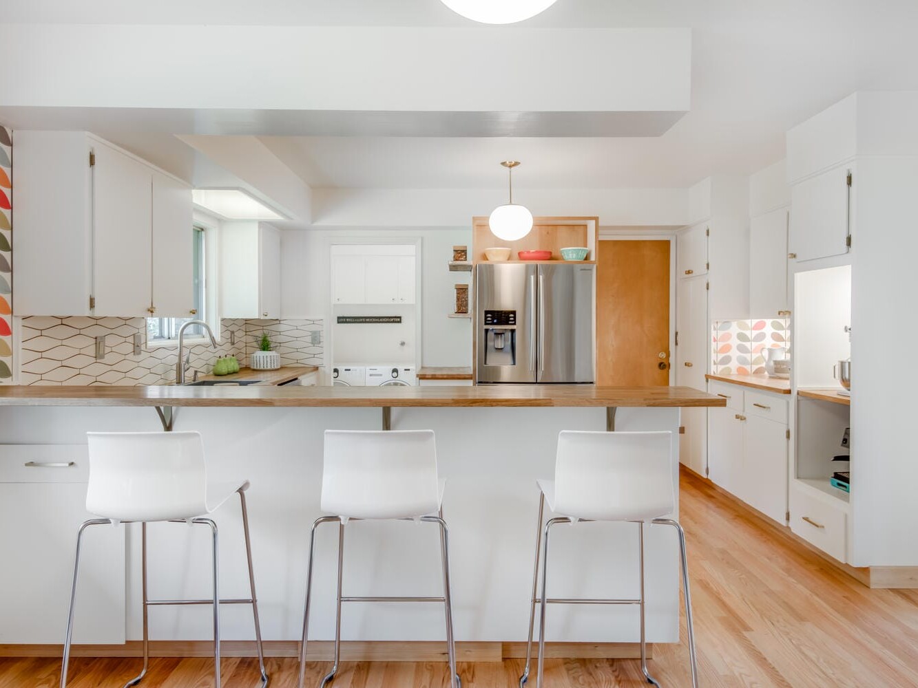 A modern kitchen in Portland, Oregon, features white cabinets, wooden flooring, and a geometric backsplash. Three white bar stools grace the island, while stainless steel appliances gleam in the background. Bright lighting and patterned wallpaper complete this stylish design.