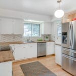 A bright kitchen in Portland, Oregon, boasts white cabinets, wooden countertops, and a stainless steel refrigerator. The geometric tile backsplash complements the colorful wallpaper on one wall, while a window above the sink floods the space with natural light. 