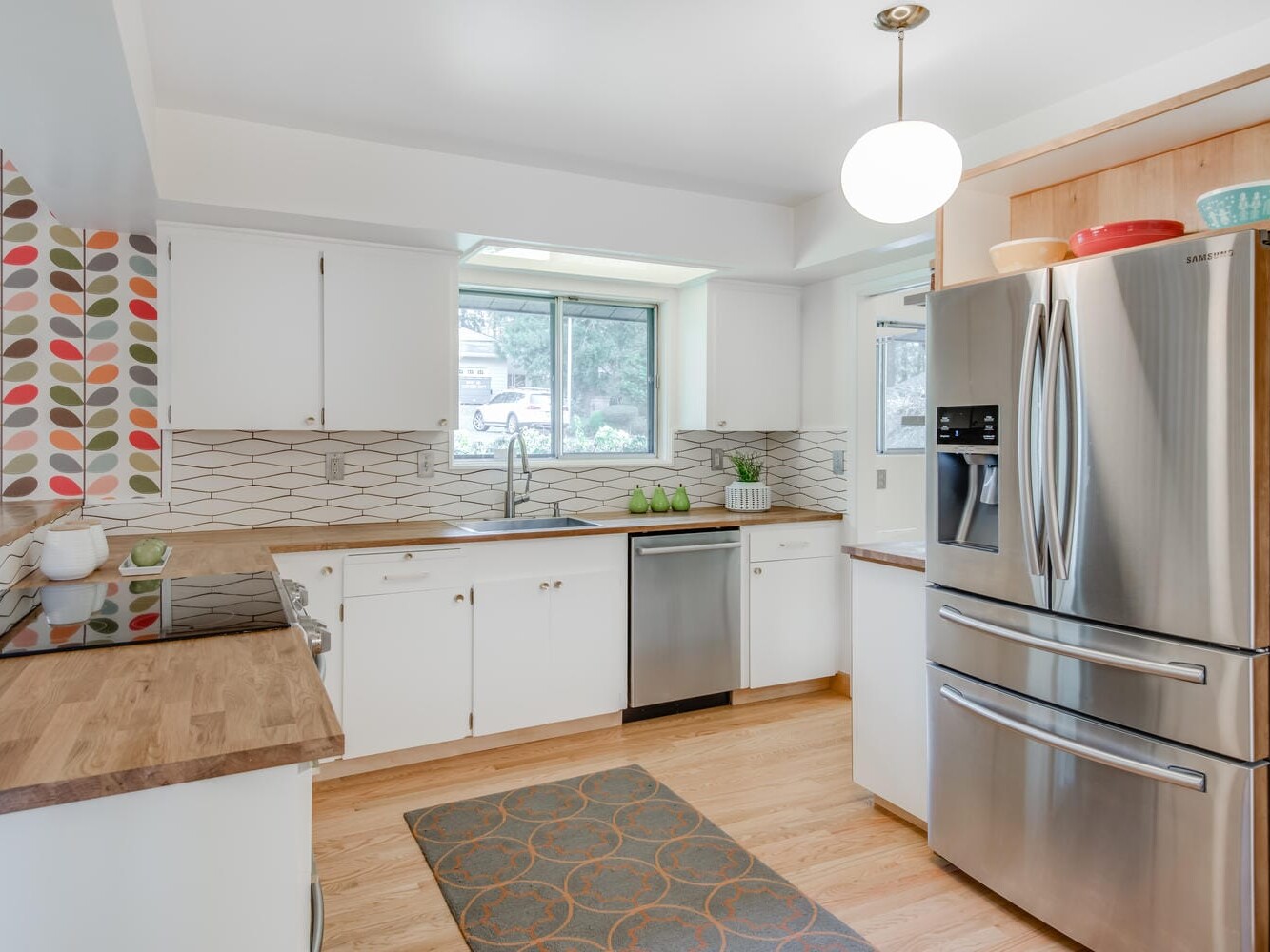 A bright kitchen in Portland, Oregon, boasts white cabinets, wooden countertops, and a stainless steel refrigerator. The geometric tile backsplash complements the colorful wallpaper on one wall, while a window above the sink floods the space with natural light.