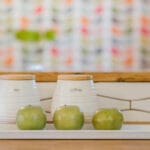 A kitchen counter in Portland, Oregon displays two white containers labeled tea and coffee, topped with wooden lids. In front, a rectangular white tray holds three green apples. The background showcases a colorful, blurred pattern. 