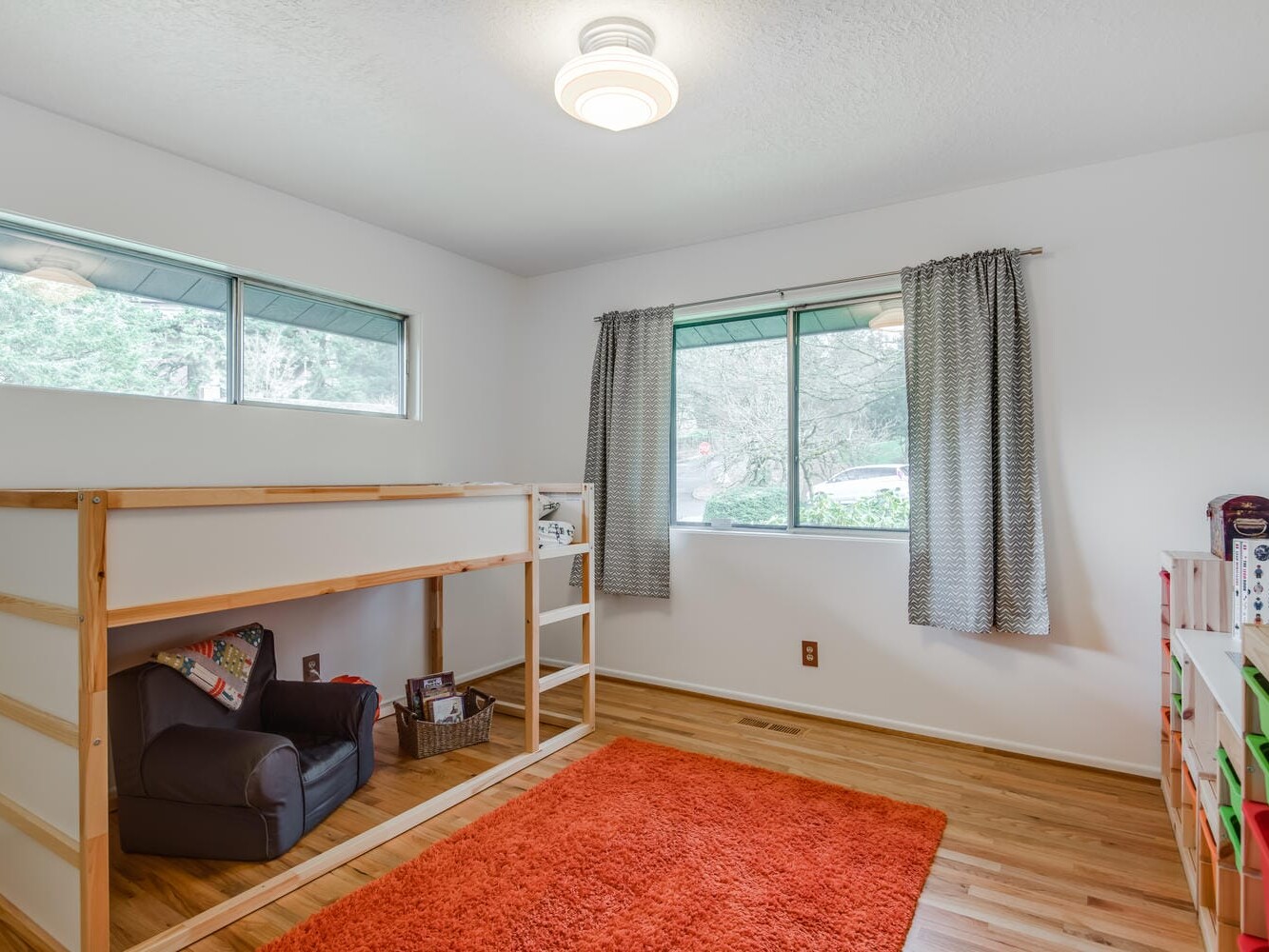 A children's room in Portland, Oregon, features a wooden loft bed with a cozy chair underneath. A bright orange rug adorns the wooden floor, with gray curtains framing the window. A colorful shelf holds books and toys against crisp white walls.