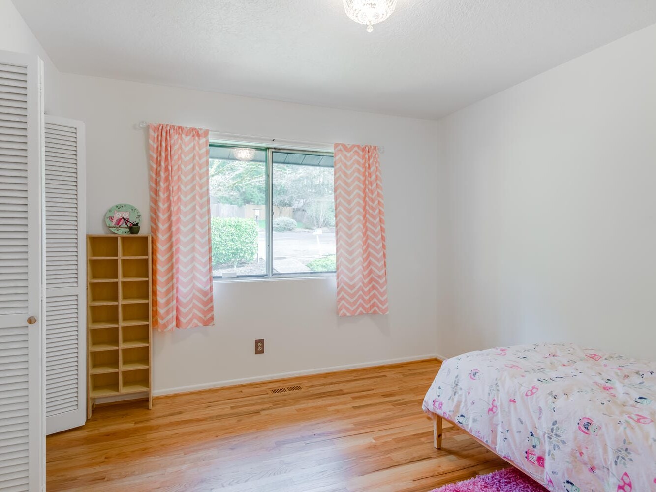 A simple bedroom in Portland, Oregon features wooden floors, a twin bed with a floral duvet, and pink chevron curtains on the window. A small wooden shelf holds a decorative plate on the left. The room is bright and minimally furnished, capturing the charm of Portland's unique style.