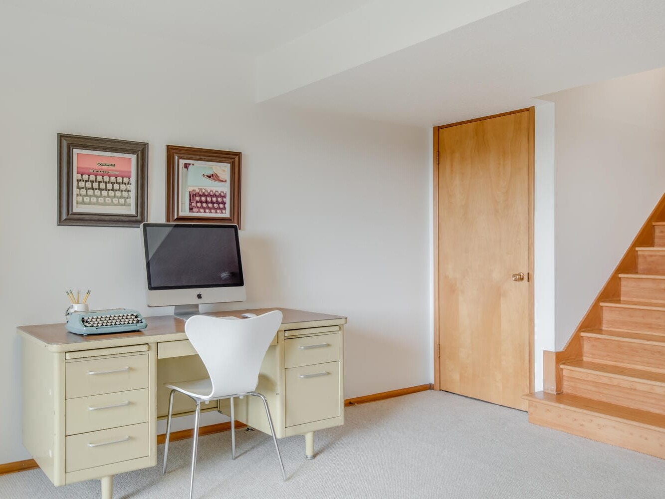 A minimalist office space in Portland, Oregon, features a beige desk with a typewriter and pencils, a computer monitor, and a white chair. Two framed pictures hang above the desk. Wooden stairs lead upwards, and the walls are painted white.