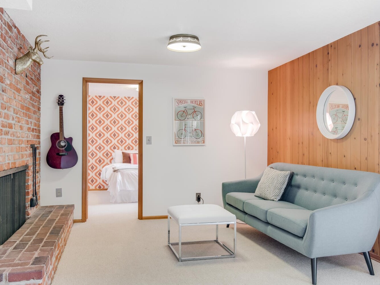 A cozy living room in Portland, Oregon, features a brick fireplace, mounted guitar, and light blue sofa. A round mirror adorns the wooden wall with a white floor lamp and square coffee table. Through the doorway lies a bedroom with patterned wallpaper, while a bicycle poster hangs above.