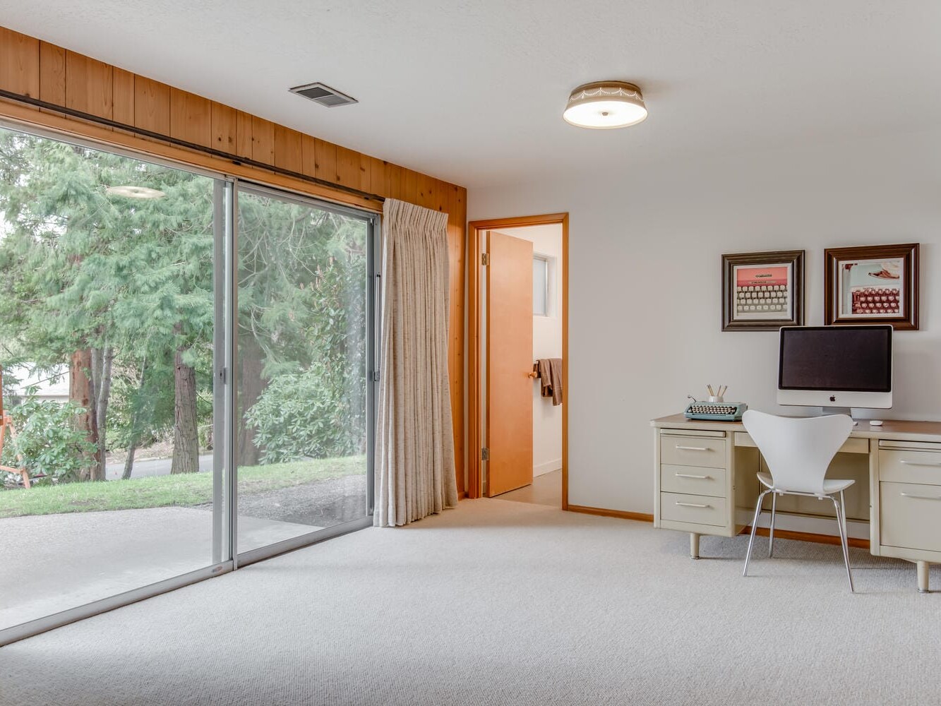 A bright Portland, Oregon room features a large sliding glass door with curtains, providing a view of trees. A desk with a computer, chair, and two framed pictures is against the wall. A doorway leads to another part of the house. The floor is carpeted.