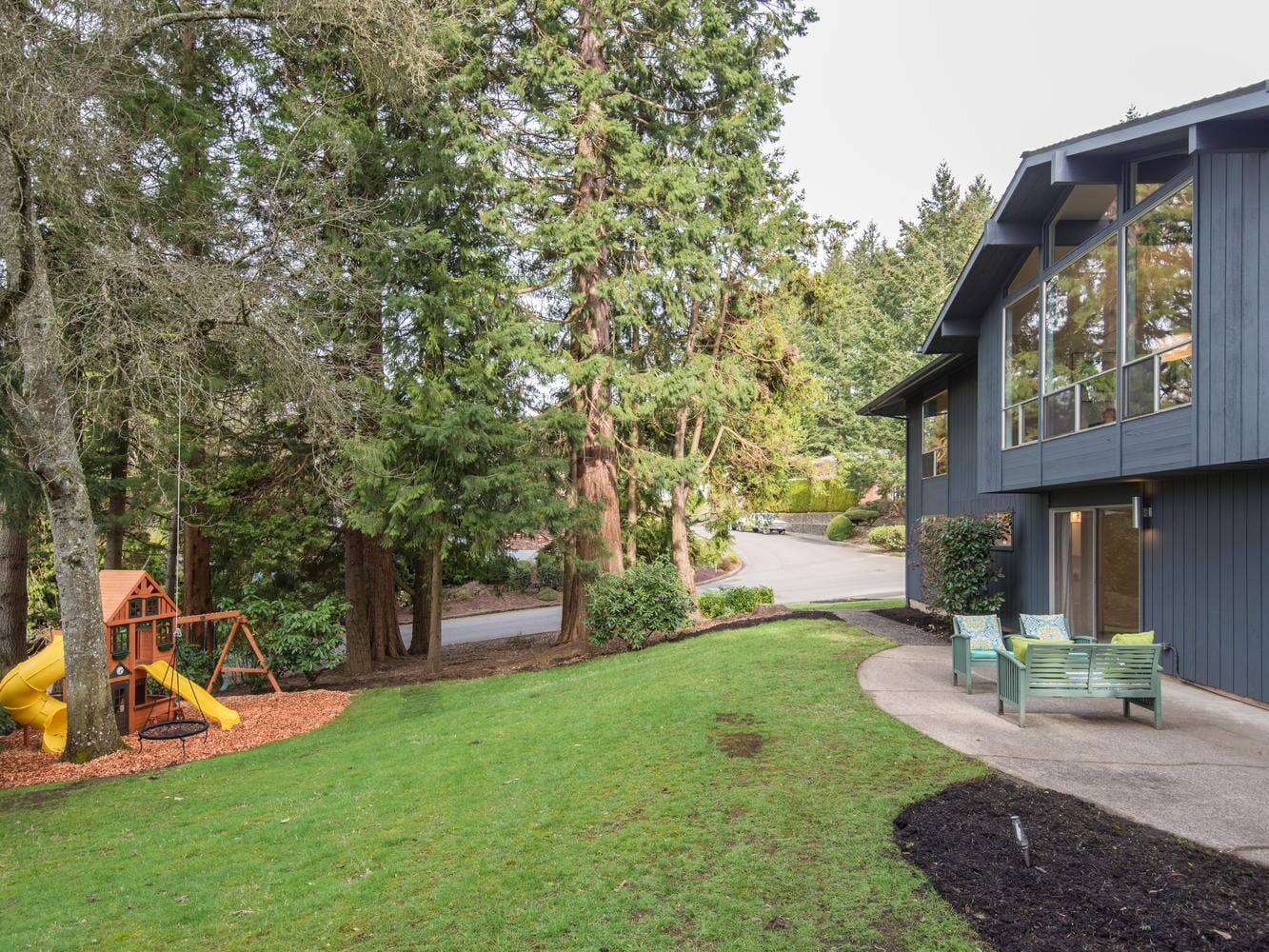 A modern blue house with large windows nestled in the tall trees of Portland, Oregon. A green lawn leads to a children's playset featuring a yellow slide. The serene, wooded neighborhood boasts a concrete path and outdoor bench alongside the house.