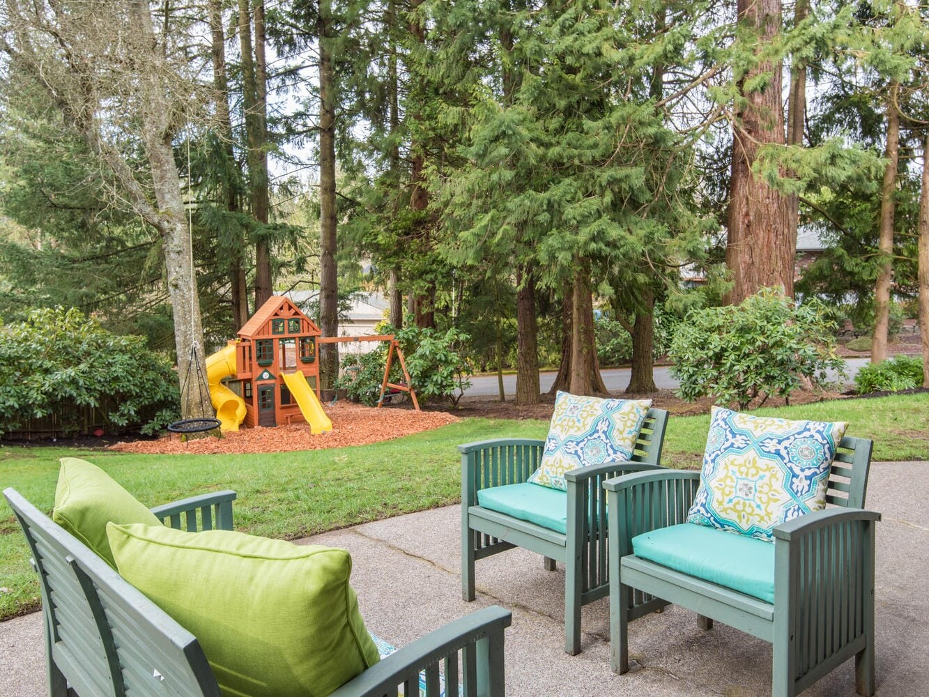 A backyard in Portland, Oregon, showcases a playset with a slide and swings nestled among tall trees. In the foreground, two wooden chairs with blue and green cushions rest on a concrete patio. The grassy area is well-maintained, reflecting the charm of this scenic city.