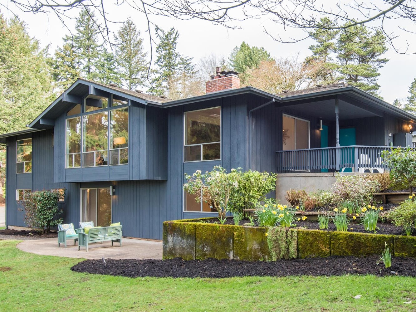 A modern two-story house with large windows and a dark exterior. It features a landscaped yard with a variety of plants and flowers, and an outdoor seating area with a green bench. There are tall trees in the background.