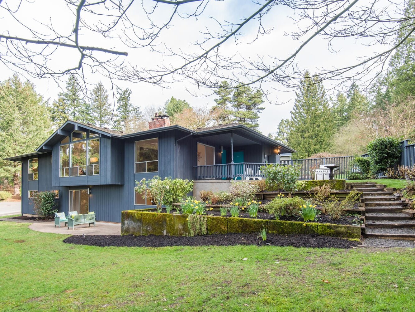 A modern, two-story house with large windows is set in a lush, green landscape. The home features dark blue exterior walls, a spacious patio, and a garden with stone steps leading to a higher terrace. Trees surround the property under a cloudy sky.