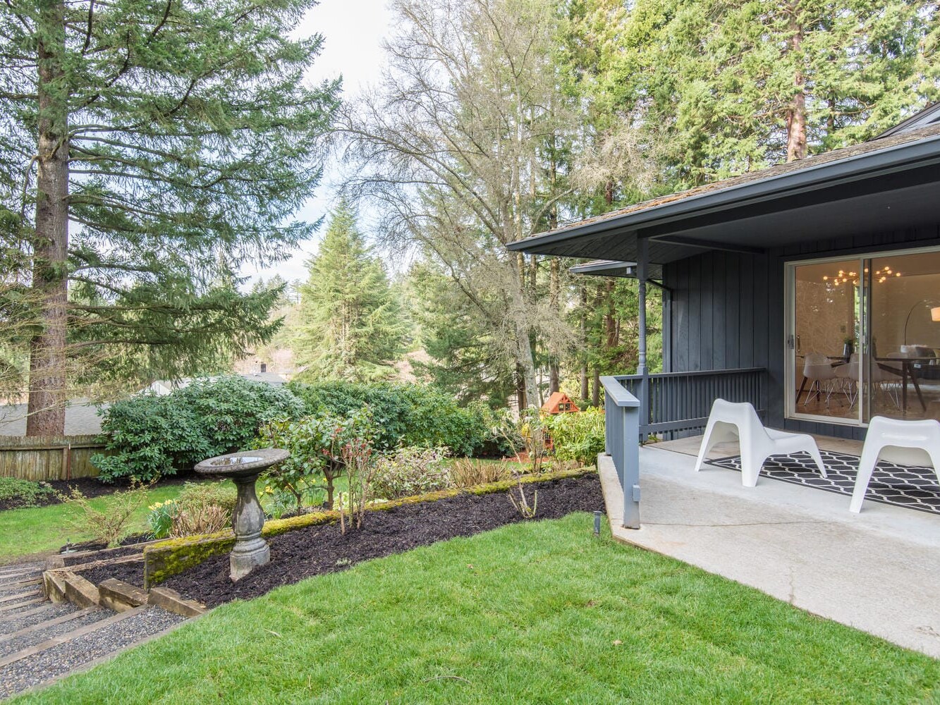 A modern house with a small porch featuring two white chairs and a patterned rug, overlooking a lush green garden. Tall trees and shrubs surround the yard, and a birdbath is seen beside wooden stairs leading downwards.
