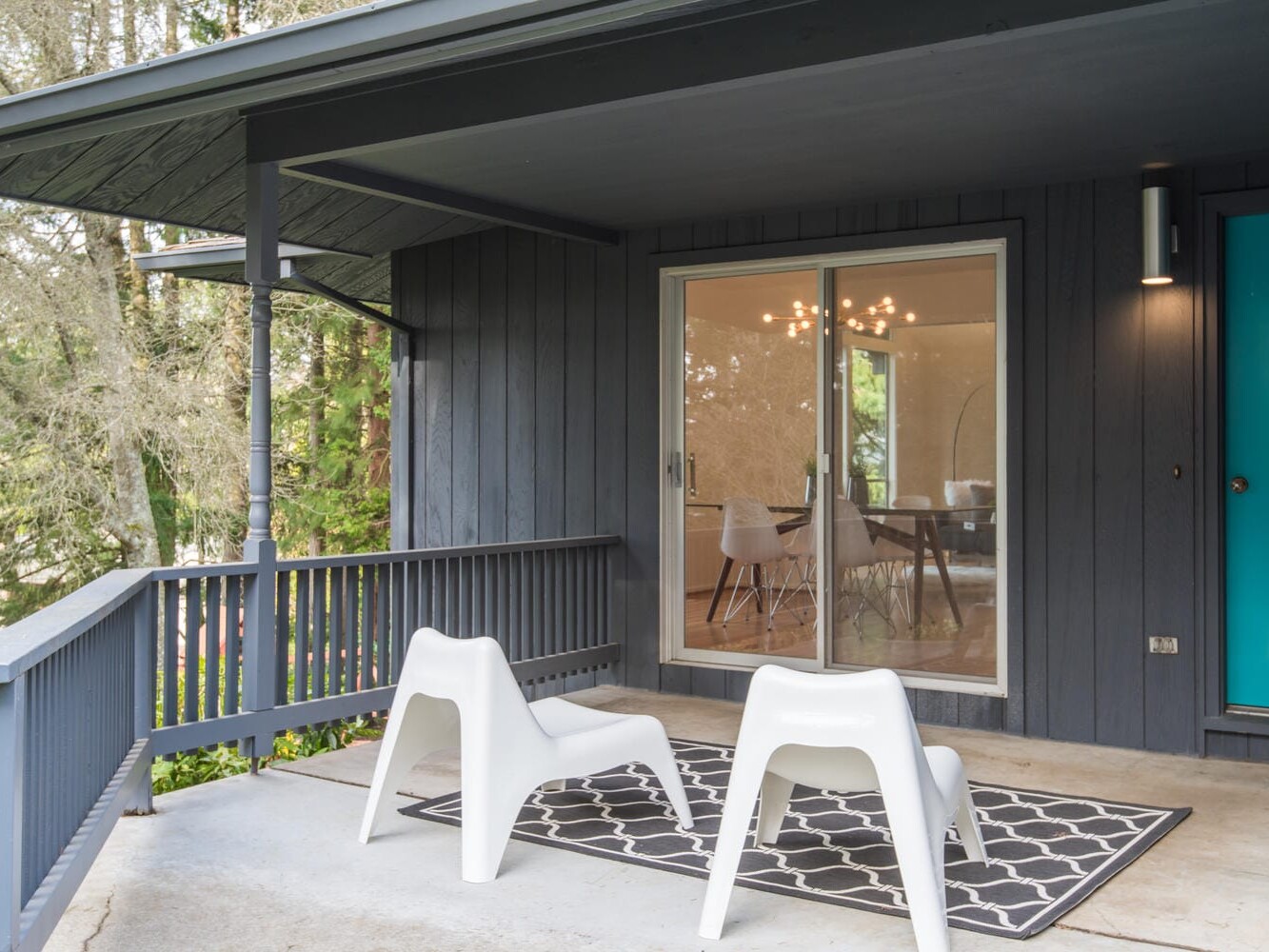 A modern porch with two white chairs on a patterned rug, facing a large sliding glass door. The exterior walls are dark gray, and the door is turquoise. Inside, a dining area is visible with a round table and overhead chandelier. Trees are in the background.