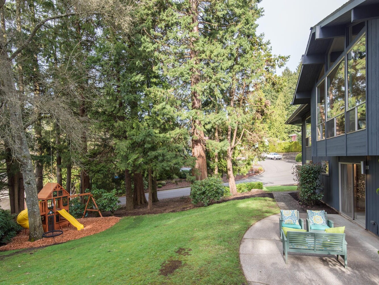 A modern house with large windows sits beside a lush green lawn. Two lounge chairs are on a patio. A wooden playset with a yellow slide is under tall trees in the background. The scene is peaceful and suburban.
