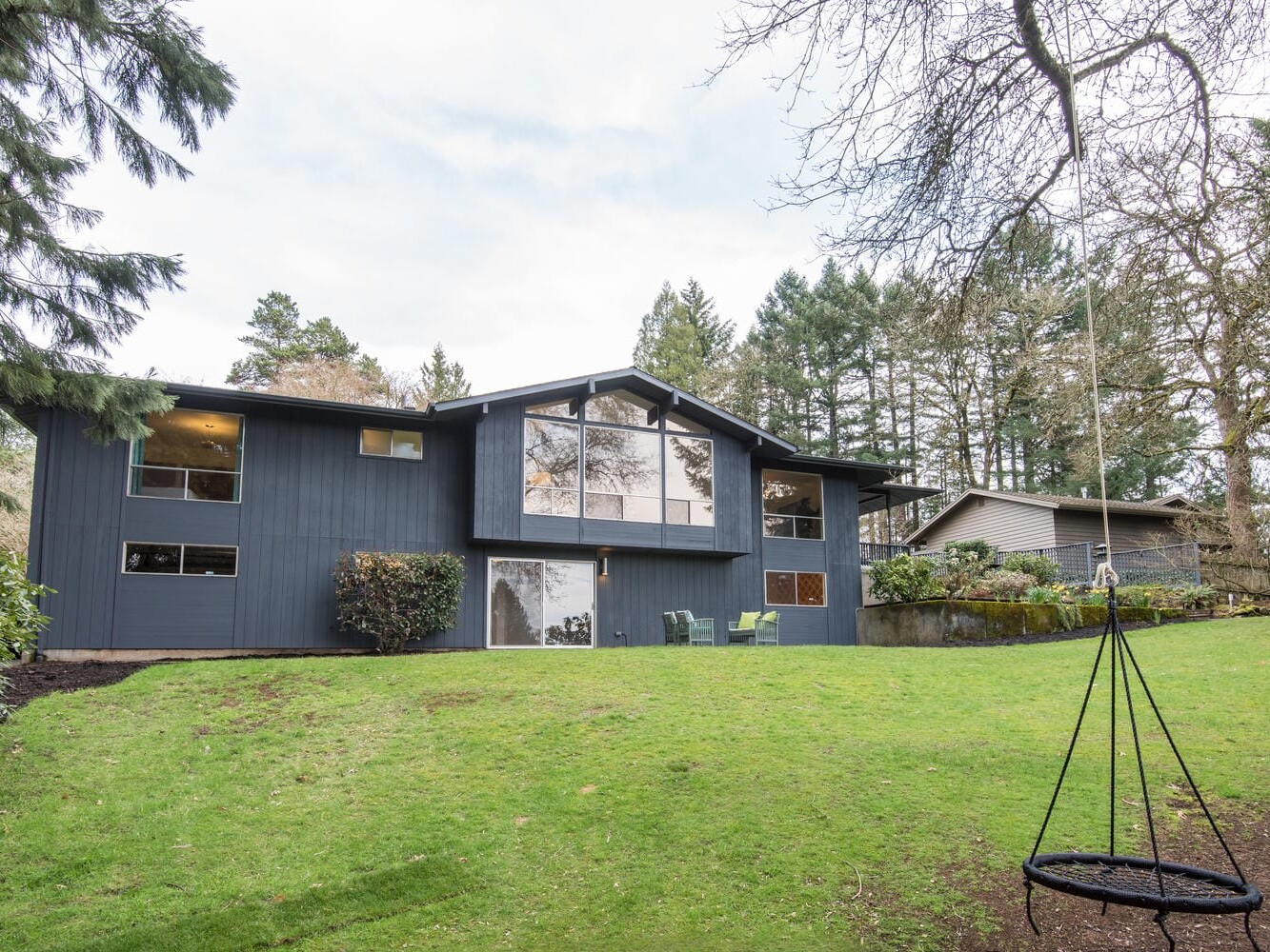 A two-story house with dark blue siding and large windows is surrounded by trees and a grassy yard. A circular swing hangs from a tree in the foreground. A neighboring house is partially visible on the right.