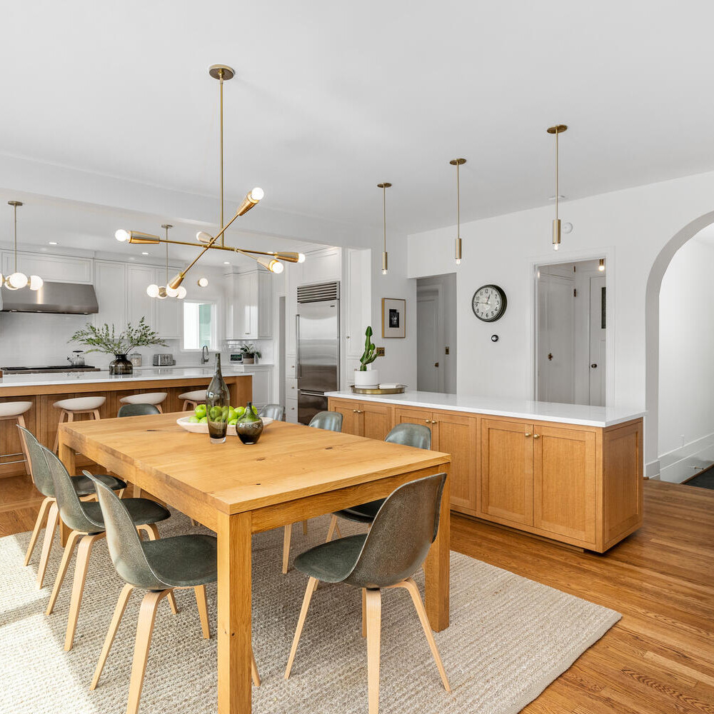 A modern kitchen and dining area in Portland, Oregon, features wooden floors and white walls. The dining table is surrounded by green chairs. The kitchen boasts a large island, white cabinetry, stainless steel appliances, and pendant lights. An arched door is visible in the background.