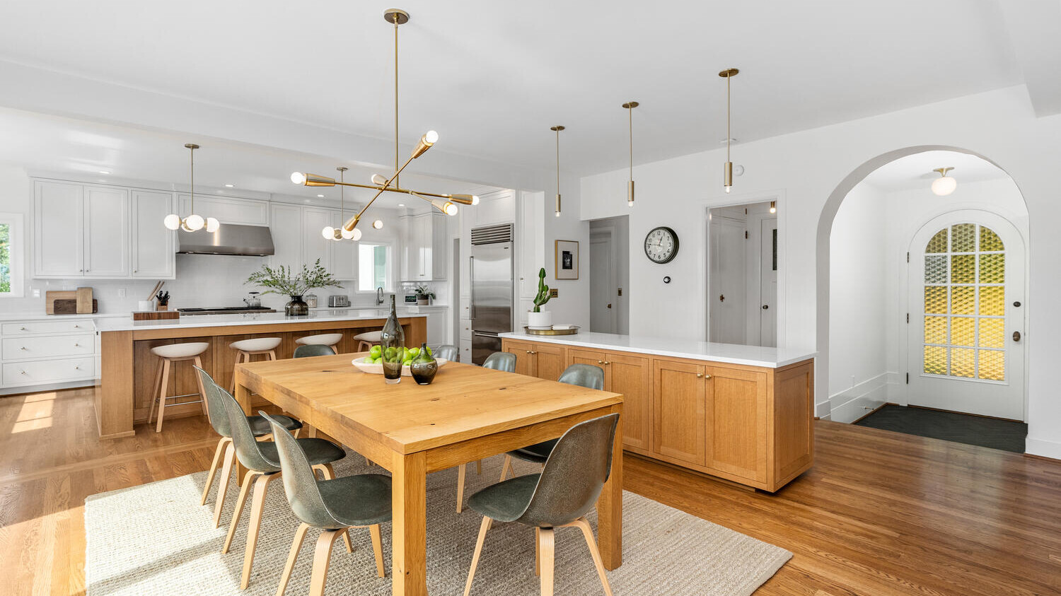 A modern kitchen and dining area in Portland, Oregon, features wooden floors and white walls. The dining table is surrounded by green chairs. The kitchen boasts a large island, white cabinetry, stainless steel appliances, and pendant lights. An arched door is visible in the background.