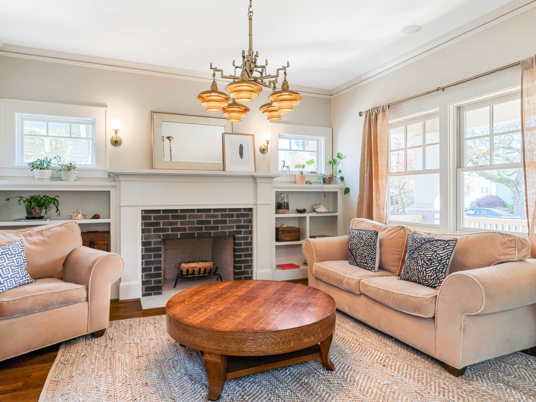A cozy Portland, Oregon living room features a brick fireplace surrounded by white built-in shelves. A beige sofa and armchair with patterned pillows sit on a light rug. A round wooden coffee table rests in the center, and a vintage chandelier hangs from the ceiling.