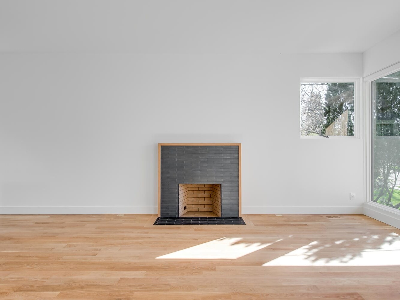A modern, minimalist room in Portland, Oregon features white walls, light wood flooring, and a small black tiled fireplace. Large windows flood the space with natural light, casting tree shadows on the floor. A lone partially visible cabinet is on the left.