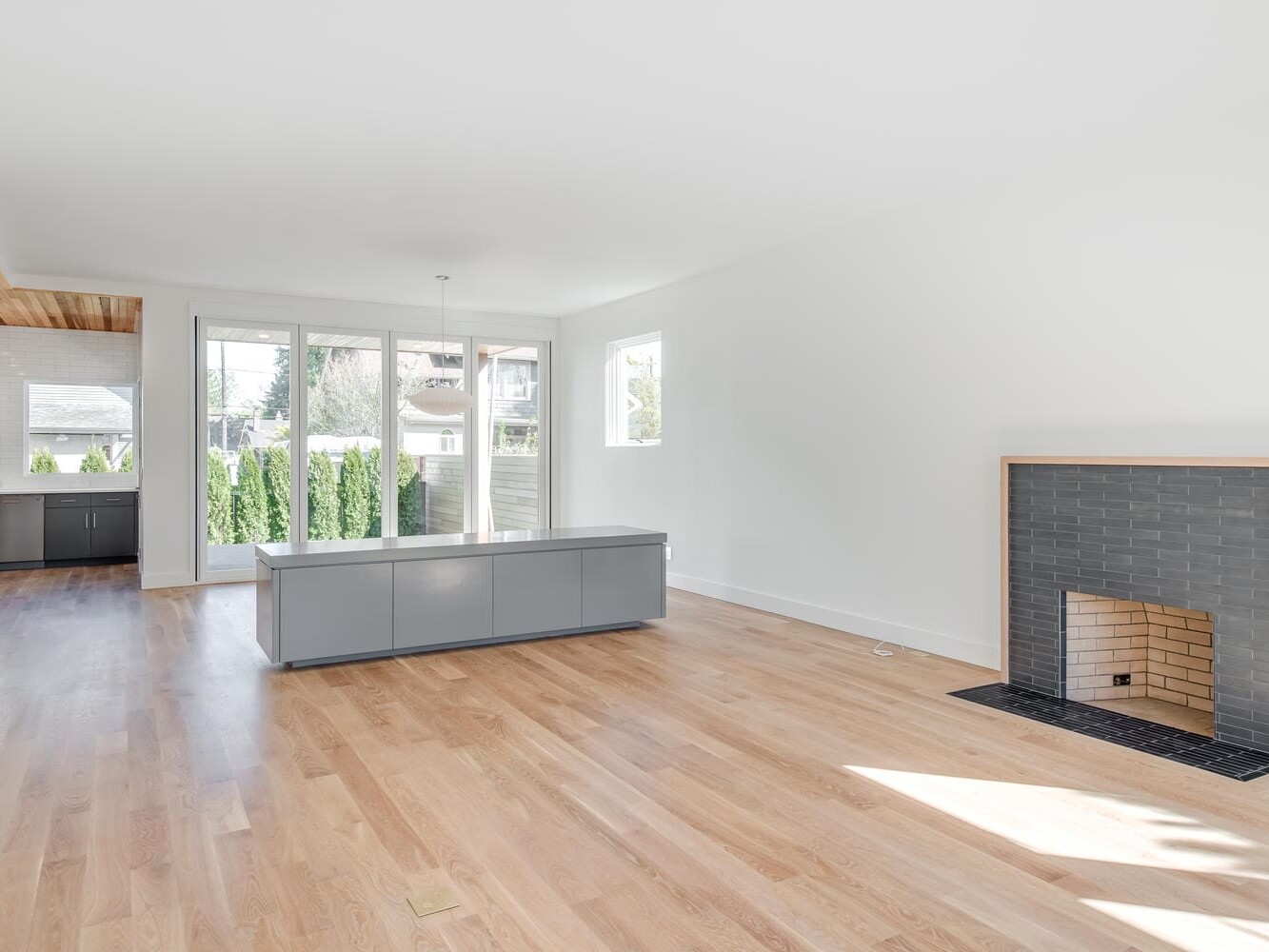 Spacious, empty living room with light wood flooring, large windows that let in Portland, Oregon's natural light, a modern gray-tiled fireplace, and an open view into a kitchen area with light gray cabinetry.