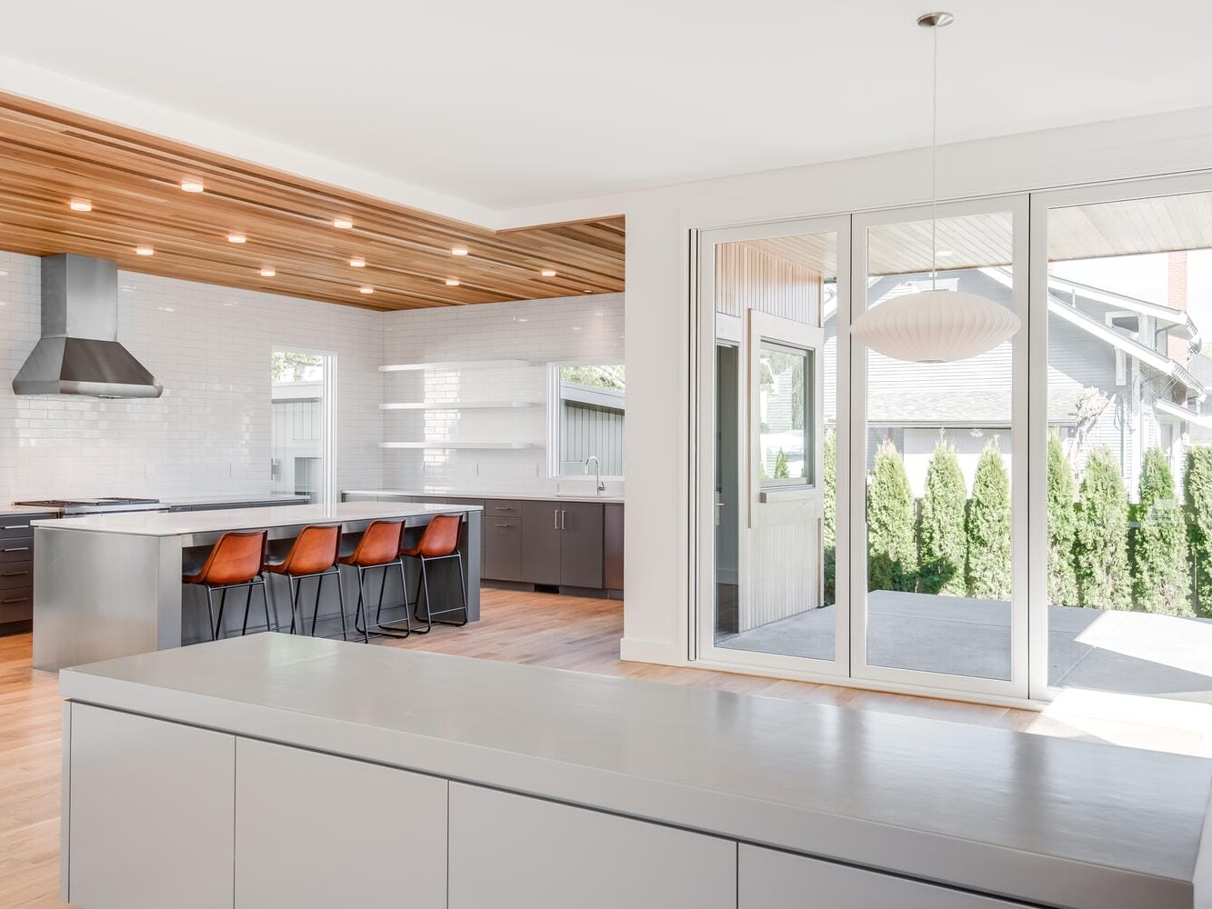 Modern kitchen in Portland, Oregon with a wooden ceiling and white walls, featuring a stainless steel range hood and an island with four brown bar stools. Sliding glass doors open to an outdoor patio with trees, allowing natural light to fill the space beautifully.
