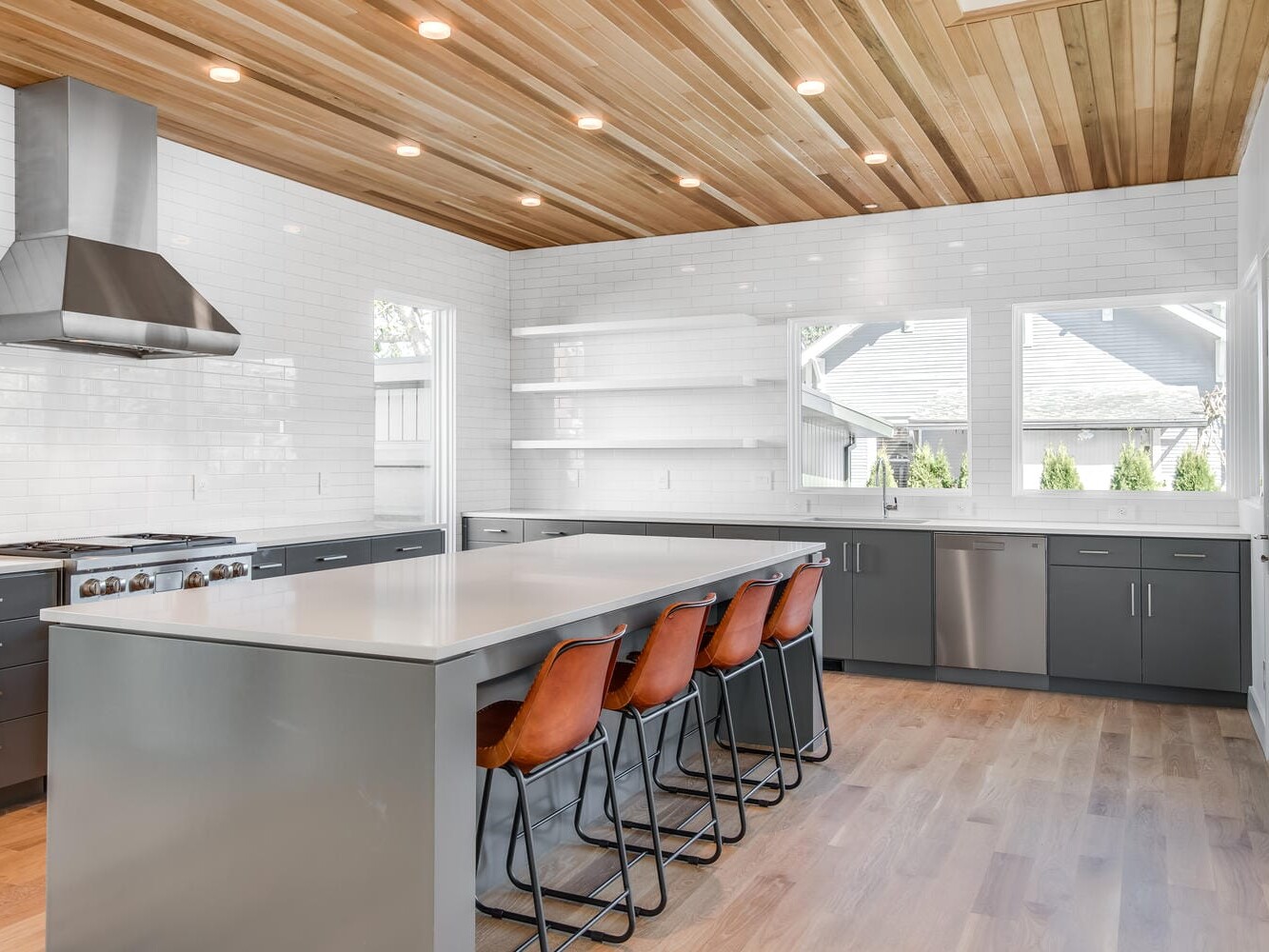 A modern kitchen in Portland, Oregon, with a wooden ceiling, recessed lighting, and light wood flooring. It features a long island with four brown chairs, gray cabinets, a stainless steel range hood, and large windows providing natural light.
