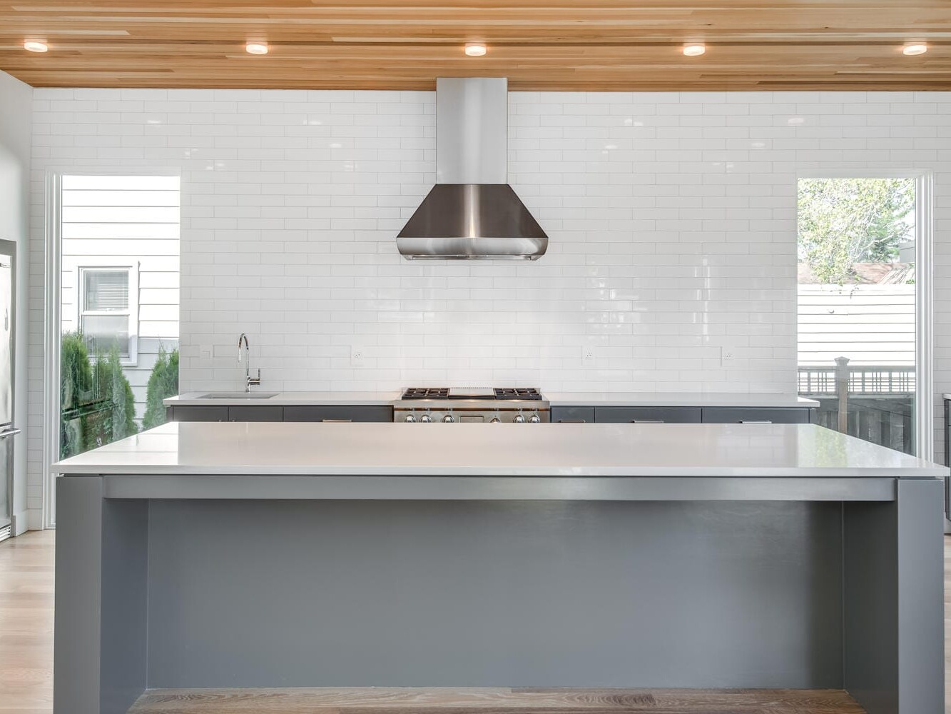 Modern kitchen in Portland, Oregon, featuring a large gray island, stainless steel appliances, and a sleek range hood. White subway tiles line the wall beneath a wooden ceiling with recessed lighting. Expansive windows invite natural light to fill the space.