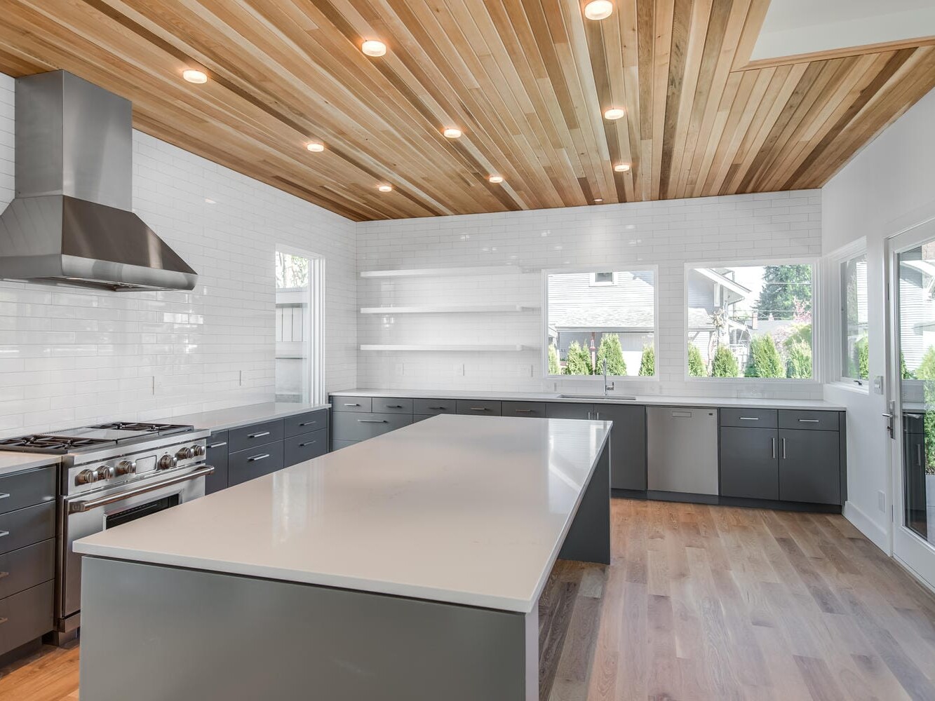 Modern Portland, Oregon kitchen with a sleek design featuring a large island with a white countertop, stainless steel appliances, dark gray cabinets, and a wooden ceiling. The space is well-lit with recessed lighting and has windows providing natural light.