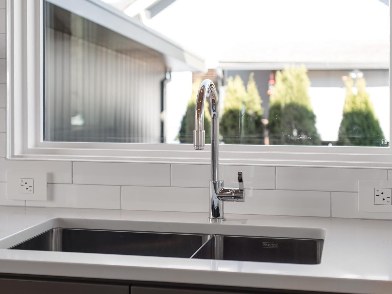 A modern kitchen sink with a stainless steel faucet is set against a backdrop of white subway tiles, reminiscent of Portland, Oregon's sleek urban design. A large window above the sink offers a view of trees and a neighboring building.