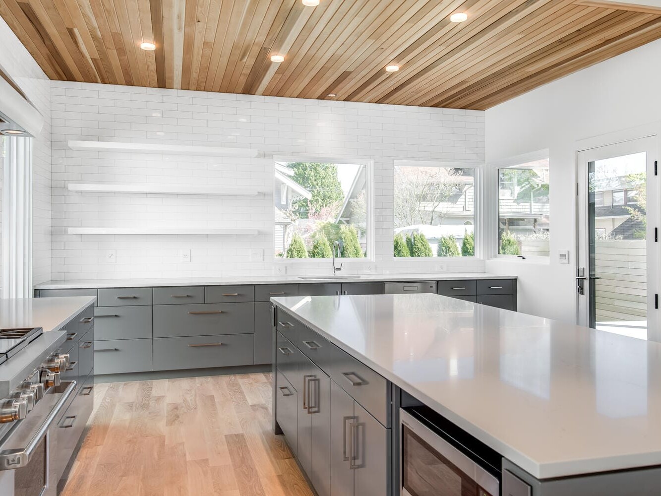 Modern kitchen in Portland, Oregon, with light wood flooring, white subway tile backsplash, and gray cabinets. A large island with a light countertop sits centrally. Windows provide natural light, and a door opens to the outdoors. Recessed lighting and open shelves complete the space.