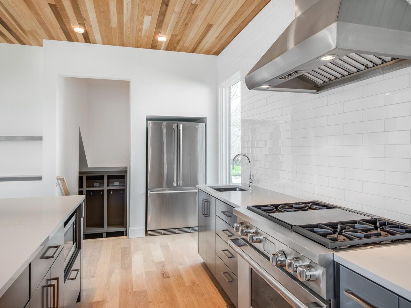 A modern kitchen in Portland, Oregon, showcases stainless steel appliances like a double-door refrigerator and a large gas stove with an overhead hood. The space is beautifully designed with wooden flooring, a wood-paneled ceiling, and crisp white tiled walls.