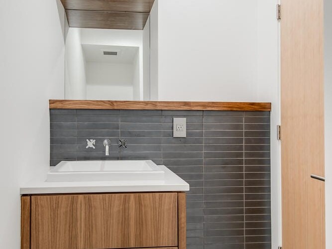 A modern bathroom in Portland, Oregon, features a floating wooden vanity and sink against a dark gray tiled wall. The floor showcases a geometric black and white pattern, while wooden floating shelves above the sink complement the light wooden door on the right.