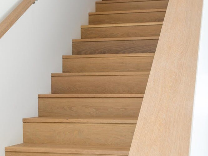 A wooden staircase with light-colored steps and a matching railing ascends between two white walls, embodying Portland Oregon's minimalist and modern design ethos. It leads up to a softly lit landing, reflecting the city's blend of natural elegance and contemporary style.