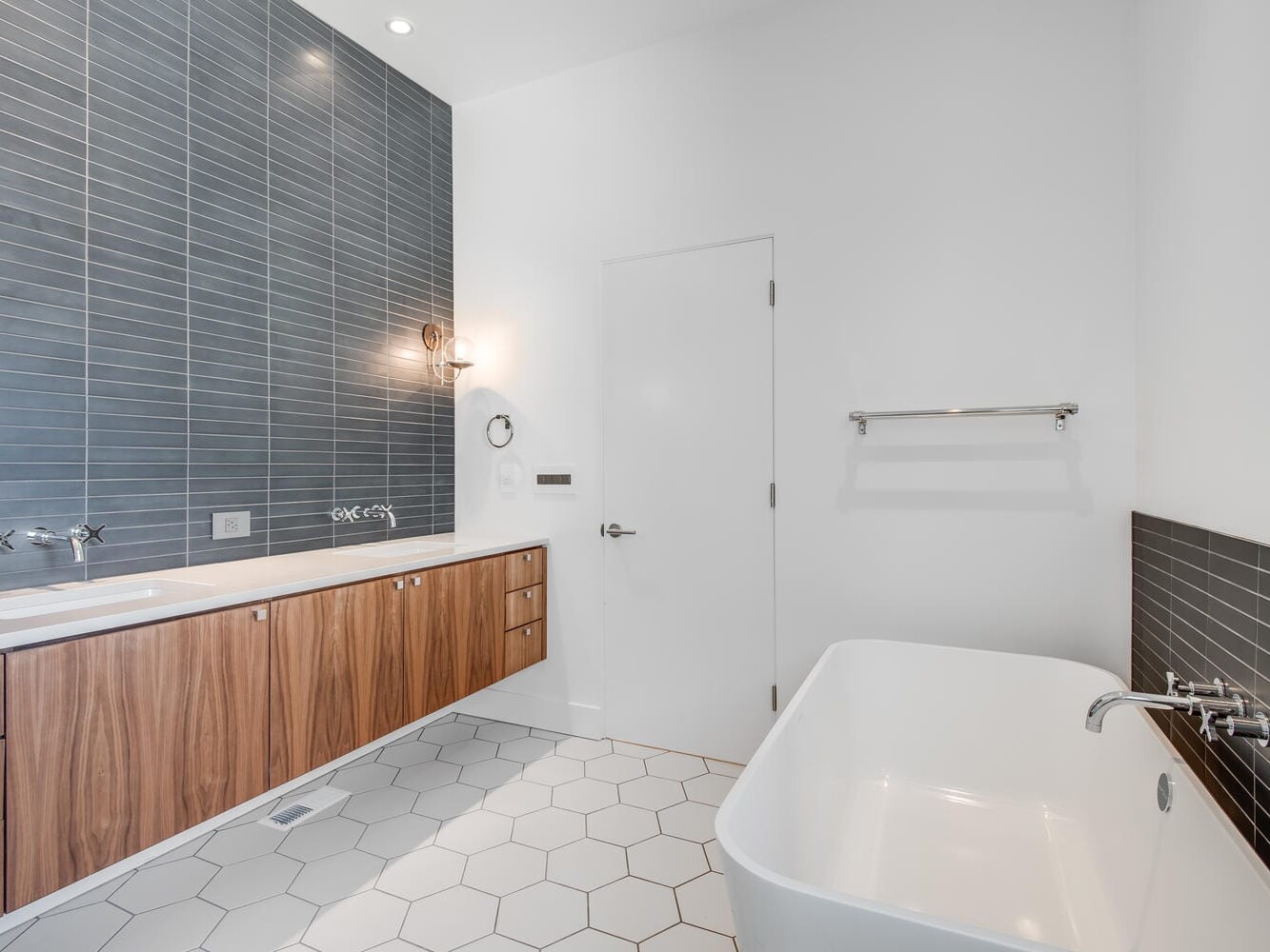 A modern bathroom in Portland, Oregon, showcases a minimalist design with a large white bathtub, hexagonal floor tiles, and a wall-mounted wooden vanity with dual sinks. The gray tiled walls are enhanced by a stylish wall sconce for lighting, complemented by a conveniently placed towel rack.