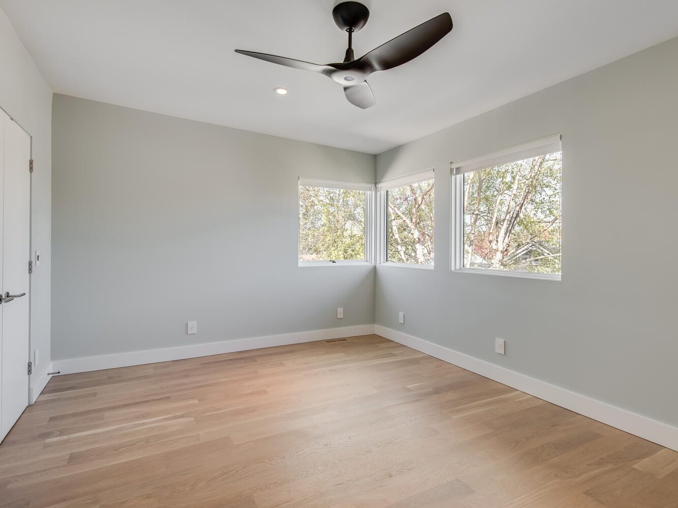 A bright, empty room with light hardwood floors, gray walls, and a black ceiling fan evokes a charming Portland, Oregon vibe. Two windows on the right side usher in natural light, revealing tree branches outside. A closed white door graces the left wall.