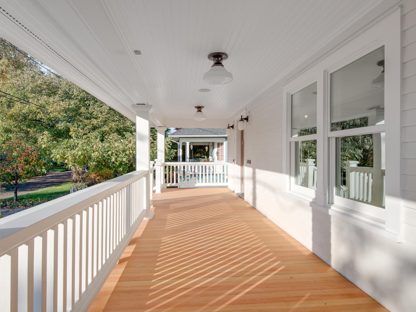 A sunlit wooden porch with white railings and ceiling, featuring two ceiling lights. The porch overlooks a green garden area, with shadows cast by the railings enhancing the cozy, inviting atmosphere.