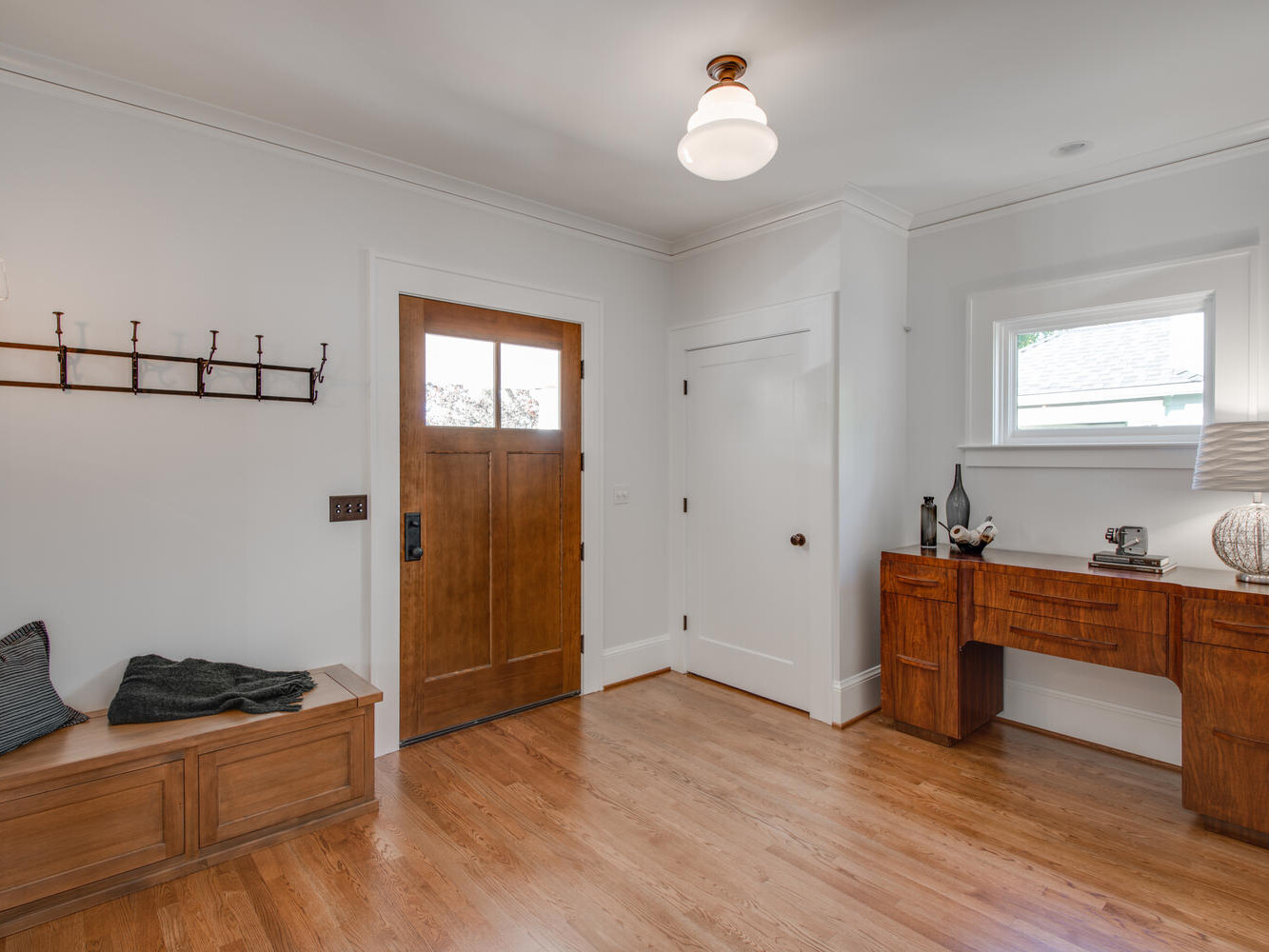A cozy entryway with a wooden bench on the left, a wooden door, and a small wooden desk with decorative items and a lamp on the right. The room has polished hardwood floors and white walls, with a ceiling light fixture.