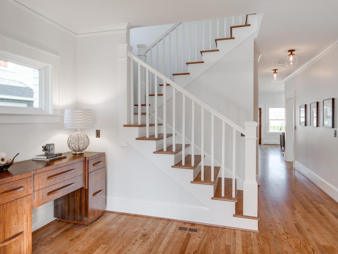 A modern interior with a wooden staircase and white railing. A wooden desk with a decorative lamp and books is situated on the left. The space has hardwood floors and framed pictures on the wall, with warm lighting from ceiling fixtures.