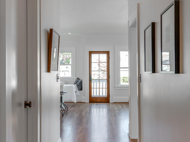 A bright hallway featuring wooden floors and white walls. Framed pictures are hung on one side, and a clear light fixture hangs from the ceiling. At the end, theres a glass-panel door leading to a room with a window and couch.
