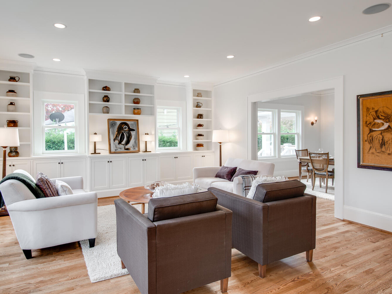 A bright and spacious living room with hardwood floors features a white sofa, two dark armchairs, and a wooden coffee table. Built-in shelves display books and decor. A window offers natural light, and a dining room is visible through an open doorway.