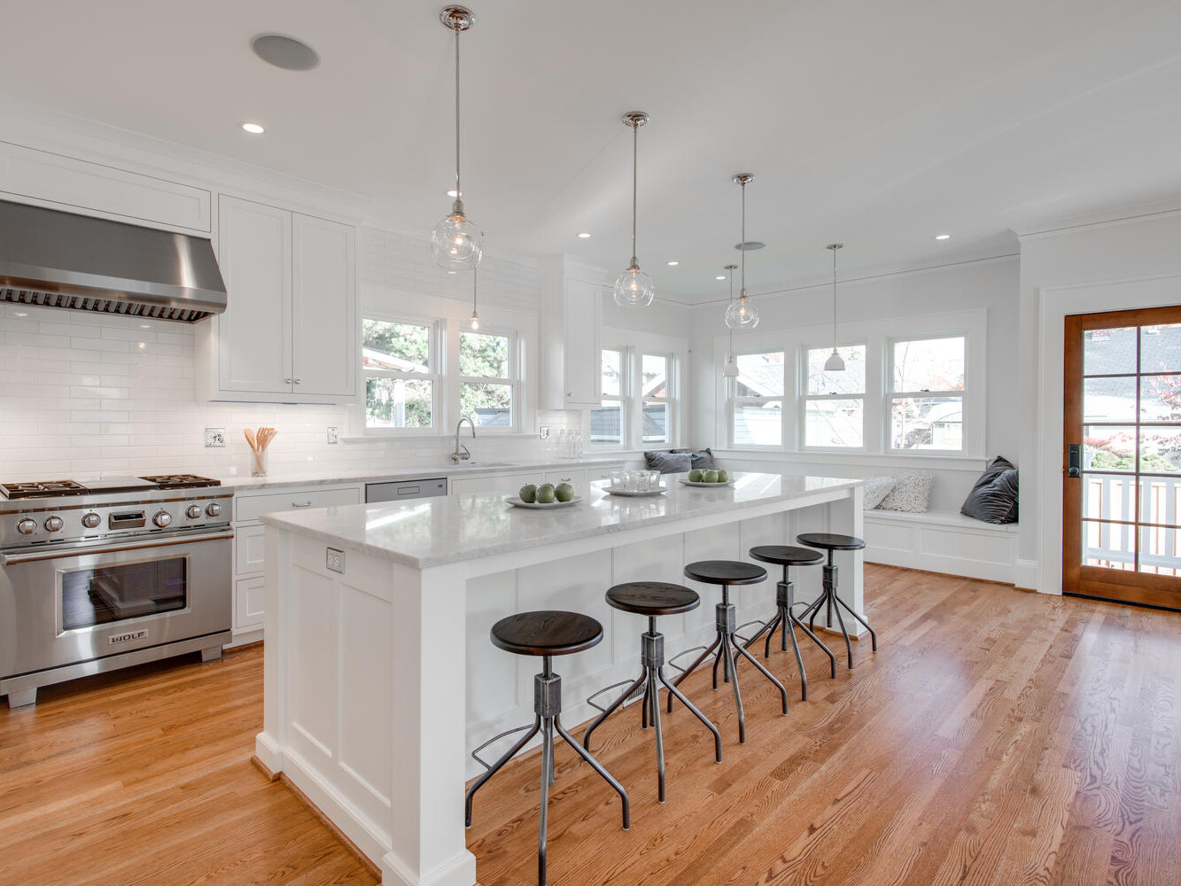 A spacious kitchen with hardwood floors, featuring a large white island and four black stools. The kitchen includes stainless steel appliances, white cabinets, pendant lights, and a wooden door leading outside. There is a cozy window seat in the corner.