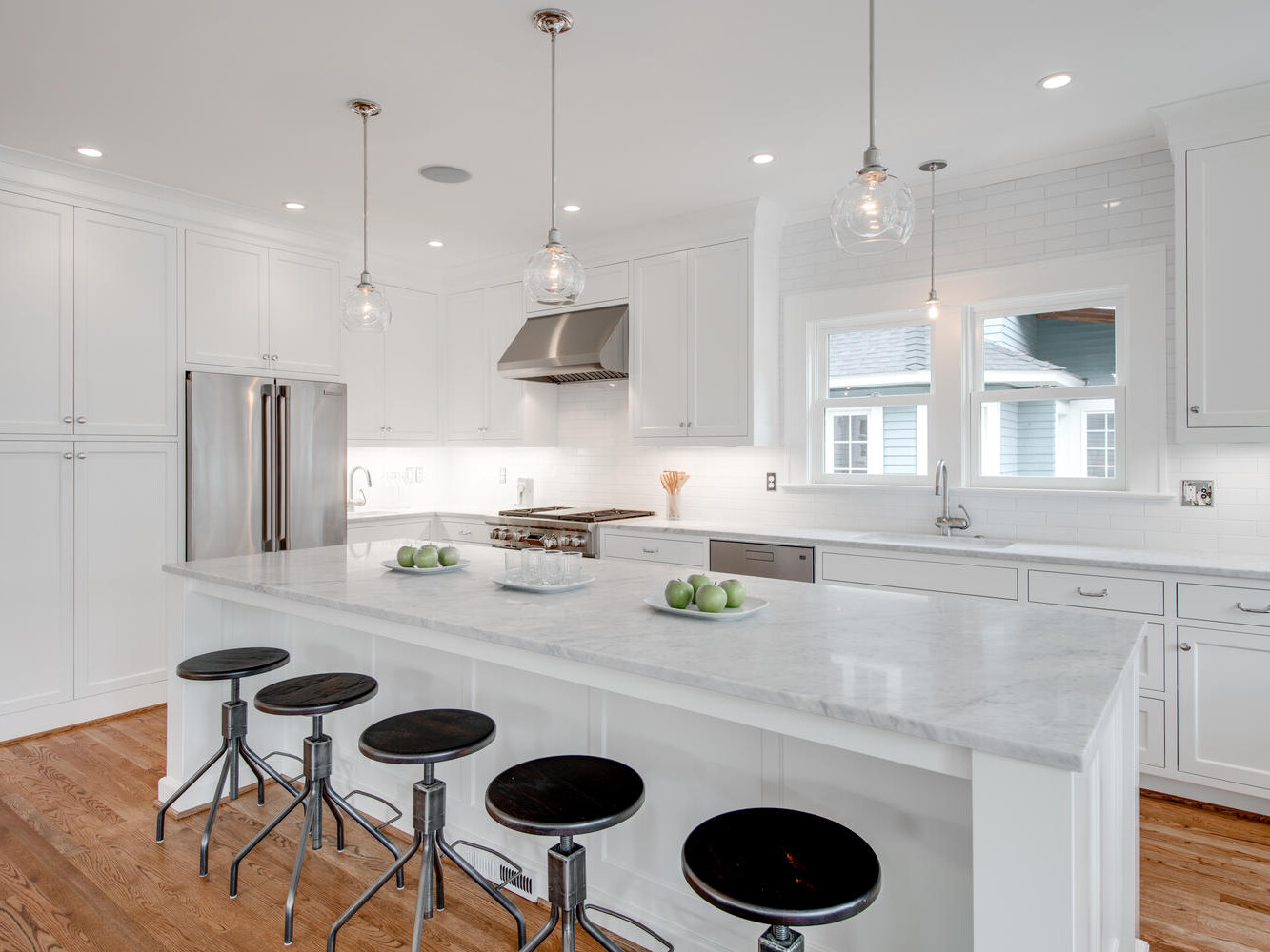 A modern kitchen with white cabinetry, marble countertops, and a central island with four black bar stools. Stainless steel appliances and pendant lights hang above the island. A bowl of green apples decorates the countertop.